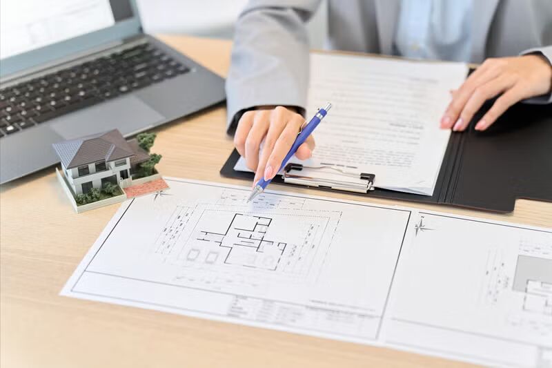 A woman analyzes a blueprint while using a laptop and writing with a pen.